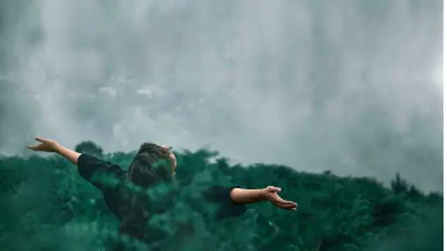 Uma pessoa parada em frente a uma parede de neblina entre plantas altas com os braços estendidos. 